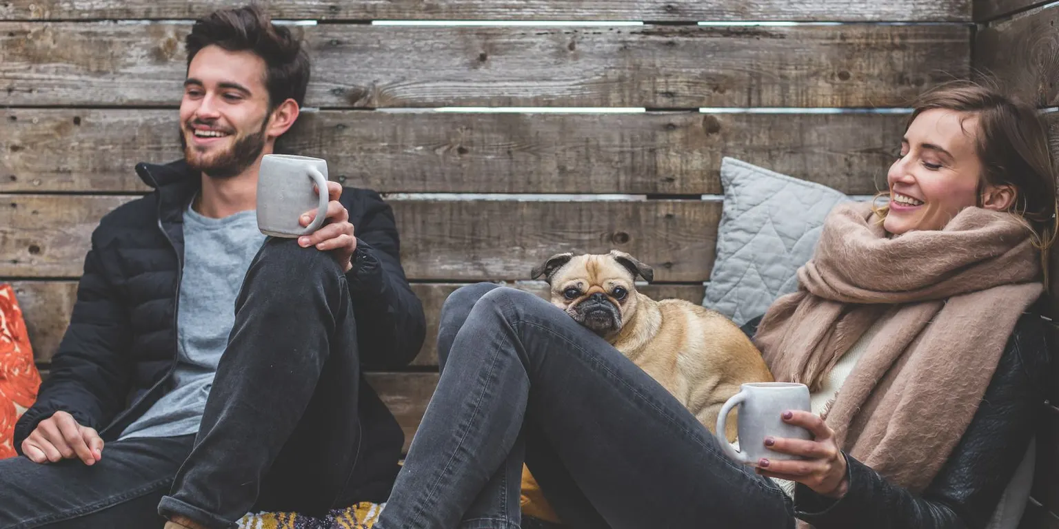 Two friends laughing over mugs of tea with a pug between them